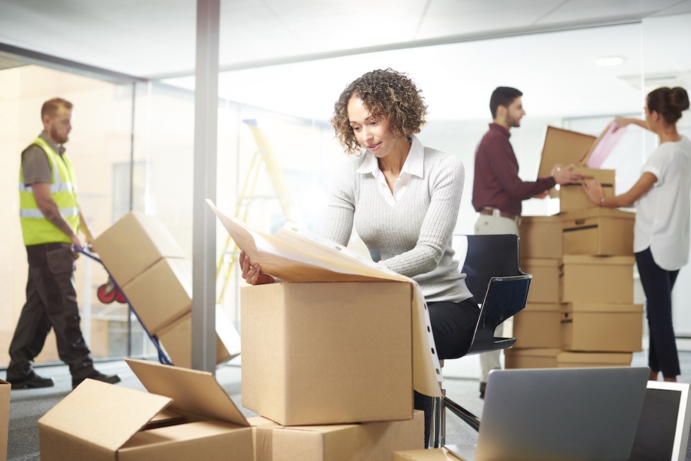 Young female office worker surrounded by document file boxes. Other workers organize files for a digital transformation of physical records.