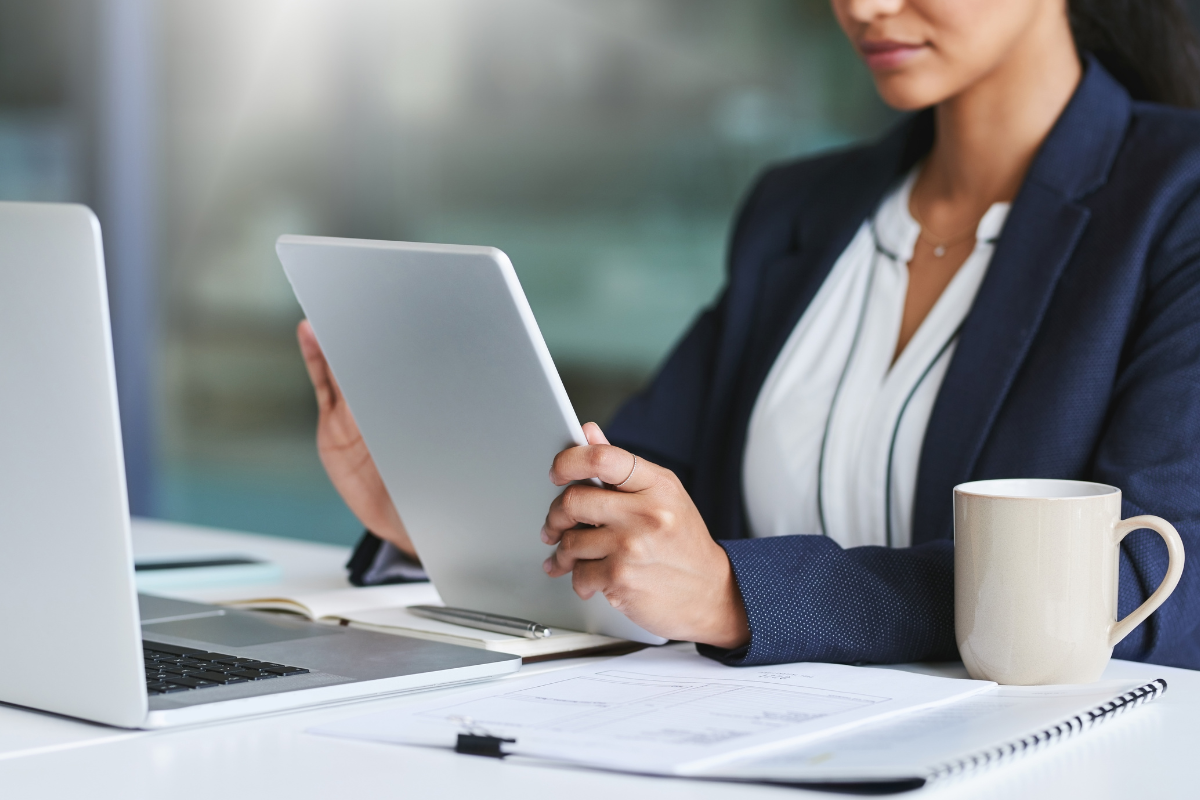 A woman reviews a file on a tablet at a desk with notes, a notepad, laptop, and mug.